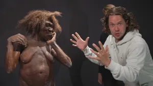 Een foto van Pascal die met zijn handen wijst naar een echt lijkend beeld van een vrouwelijk oermens van de soort Homo erectus. De vrouw is naakt en heeft een bos bruin haar en een huid met zanderige vlekken. Ze krabt met twee vingers op haar wang.