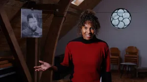 Een foto van een presentator in een rode trui met zwarte randen bij de nek en armen. Op de achtergrond hangt een zwarte lijn met een zwart-wit foto van Nelson Mandela eraan. De presentator staat op een zolder met een rond raam met een bloemvorm erin. 