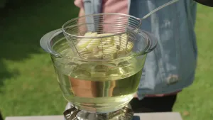 Een foto waarin iemand een klein mandje met patat in een glazen kom met olie doet. Onder de kom staat een gasbrander. De foto is genomen buiten op een grasveld. Van de persoon op de foto is geen gezicht te zien, alleen een gedeelte van de kleding.