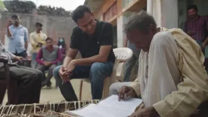 Een foto van presentator Narsingh die op een witte plastic stoel zit in een dorp in India. Naast hem zit een oudere man op een bed en wijst op een geschreven tekstregel in het opengeklapte boek voor zich. Op de foto kijken nog zeven andere mensen toe.