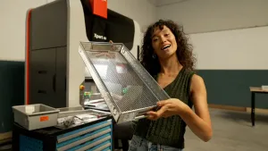 Een foto van een presentator die een stalen mand omhoog houdt die van gebruikte medische instrumenten gemaakt is.  Op de achtergrond is een werkruimte met een grote machine zichtbaar. Op de machine is de tekst "Xpert 40" te lezen.