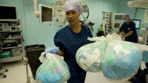 Een foto van een presentator in de operatiekamer van een ziekenhuis. De presentator heeft drie doorzichtige vuilniszakken met afval in handen. De presentator draagt kleding die artsen tijdens het uitvoeren van een operatie dragen.  