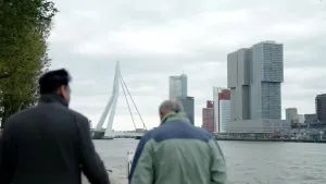Een foto van twee mannen die op de kade lopen. Op de achtergrond is de Erasmusbrug te zien. Dat is een bekende brug in Rotterdam.