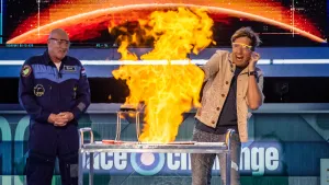 Een foto van de presentator en André Kuipers die in de Space Challenge studio staan met veiligheidsbrillen op. Voor hun staat een tafel waar een grote vlam vanaf komt door een brandbare vloeistof. 