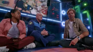 Een foto van de presentator, André Kuipers en een meisje die naast op de grond zitten in de Space Challenge studio. Ze zitten in een tent waar je doorheen kan kijken.