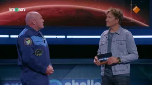 Een foto van astronaut André Kuipers en een presentator die voor een beeldscherm staan. Op het scherm is de bovenkant van de "rode planeet" Mars te zien. André heeft zijn handen gevouwen voor zijn buik en draagt een blauw astronautenpak met drie logo's.