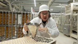Een foto van Pascal in een dropfabriek in een witte jas, witte pet, met bril en haarnetje. In zijn handen heeft hij een stalen trechter, waar zwarte vloeistof uitloopt. De vloeistof loopt in een mal waar allemaal gaatjes inziten in de vorm van een dropje.