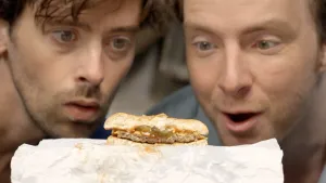 Een foto van presentatoren Willem en Pieter die met hun gezichten dicht op een hamburger die dwars door midden is gesneden en bovenop de papieren verpakking ligt.