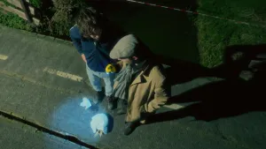 Een foto van presentatoren Willem en Pieter die buiten in het donker staan. Pieter schijnt met een zaklamp op twee stukken doek die op de stoep liggen. Onder het grootste stuk doek steken de poten van een kip uit.