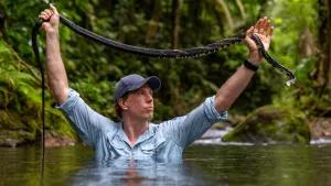 Een foto van presentator Freek die tot zijn borst in het water staat en met twee handen boven zijn hoofd een slang vasthoudt.