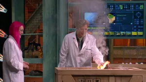 Een foto van een man en een vrouw die in de Earth Challenge studio staan, beide hebben een laboratorium jas aan. De man doet een proef met vuur bovenop een grote kist.