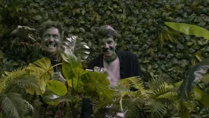 Een foto van de presentatoren die verstopt zijn door groene planten voor hun en een muur van bladeren achter hun ruggen. Ook hun gezichten zijn groen.