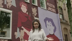 Een foto van presentator Eva die voor een muur van een huis staat met schilderingen en de naam van Aletta Jacobs en "dr.", dat betekend dokter. Aletta is met rode trui op een lichtpaarse achtergrond te zien en in een rode jurk naast een vorm van een man.