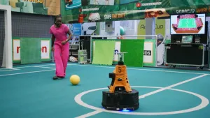 Een foto van presentator Tirsa die binnen op een voetbalveld staat en klaar staat om een gele bal te trappen. In het midden van het veld staat er een robot. Achterin bij de muur staat er een laptop met een grote televisie erboven die het veld laat zien.