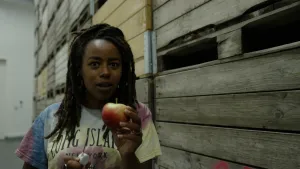 Een foto van presentator Eva die voor de camera naast een hoop opgestapelde houten kisten staat met een appel in haar hand.