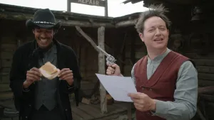 Een foto van twee mannen. De voorste heeft een blad papier in zijn hand en een pistool in zijn andere hand. De andere man draagt een cowboyhoed en heeft een broodje pindakaas in zijn handen. Op de achtergrond is er deel van een houten gebouw te zien.