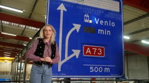 Een foto van presentator Janouk die naast een grote blauwe snelweg verkeersbord staat waar een afslag naar Venlo en een andere snelweg op wordt aangegeven.