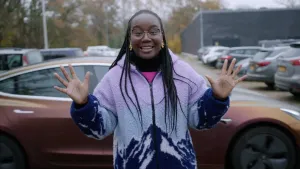 Een foto van presentator Tirsa die op een parkeerplaats voor een zelfrijdende auto staat.