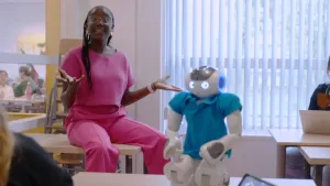 Een foto van presentator Tirsa die in een klaslokaal op een tafel zit. Op de voorgrond staat er een robot op een tafel. De robot heeft een blauw shirt aan.