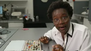 Een foto van een presentator die voor en tafel zit. Op de tafel staat een doos met edelstenen die op een zachte ondergrond liggen. Het zijn allemaal kleine edelstenen van heel diverse kleuren.