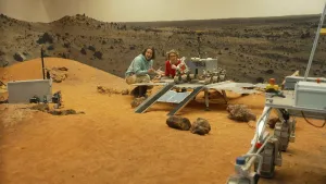 Een foto van een als de planeet Mars ingerichte ruimte met een kale heuvel van zand met enkele kleine stenen erop. Op de heuvel is  een robotwagen van Esa uitgeklapt te zien. Achter de wagen zitten de presentator en Planeetwetenschapper Inge Loes. 