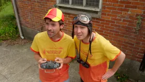 Een foto van presentatoren Willem en Pieter die buiten voor een huis staan. Willem houdt een afstandsbediening met joysticks vast die een afstand bestuurbare vliegtuig bestuurd.