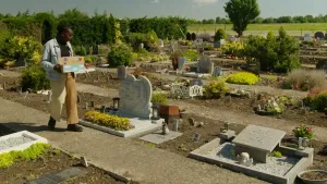 Een foto van een begraafplaats. De presentator loopt over een pad met een kleurige doos in haar hand. Ze kijkt naar een grafsteen.