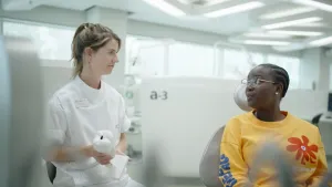 Een foto van twee mensen in een lichte ruimte. Een persoon heeft witte tandartskleren aan en heeft een plastic versie van een kies met een gaatje in haar hand. De ander zit op de tandartsstoel.