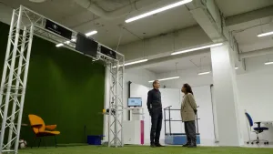 Een foto van twee mensen die staan te praten in een grote ruimte. Op de grond en tegen een muur zit kunstgras. Er hangt een constructie van metalen buizen boven het kunstgras, waar een scherm aan hangt.