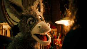 Een foto van ezel Chiel die vlak voor de camera staat. Op de achtergrond is een donkere kamer te zien waar een lamp brandt die bovenop een tafel staat met allerlei spullen erop.