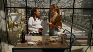 Een foto van een presentator en een mevrouw achter een kar in een grote tuinkas. Op de kar staan een mixer, een bus poedersuiker, een zak hazelnoten, twee borden met fijngemalen poeder, een bakje noten, een gesneden wit bolletje brood en een weegschaal. 