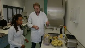 Een foto van een presentator die op een kruk zit en een man die naast de presentator staat voor een aanrecht in een laboratorium. Voor de presentator staat een plastic bak met aardappelschillen en daarachter een bord met geschilde aardappelen.