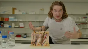 Een foto van presentator Pascal die achter een tafel in een laboratorium staat. Op tafel staat een model van de huid waarbij de huid van binnen te zien is, met bloedvaten, haren en haarzakjes.