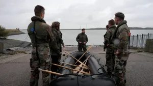 Een foto van presentator Pascal die samen met vier mariniers naast een opblaasboot staat waar roeispanen opliggen. Ze krijgen uitleg van een andere marinier die voor de opblaasboot staat. Op de achtergrond is een watervlakte te zien.