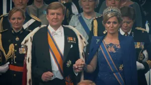 Een foto van Koning Willem-Alexander en Koningin Máxima die elkaars hand vasthouden. Koning Willem-Alexander draagt een koningsmantel. Op de achtergrond is er publiek zichtbaar. 