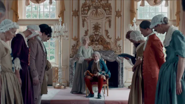 Een foto van twee mensen van adel die een witte pruik dragen: een man die op een stoel voor de open haard zit en een vrouw die achter de stoel staat. Op de voorgrond staan links en rechts een rij mensen met licht gebogen hoofd.