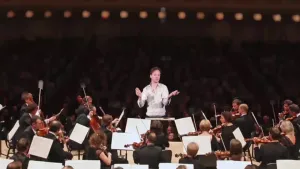 Een foto van een presentator die op de positie van de dirigent staat voor een orkest dat op een podium muziek speelt.