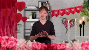 Een foto van een presentator in een romantisch ingerichte huiskamer. Vooraan zijn roze en rode rozen te zien, er hangen harten van rood papier in de achtergrond en er staan brandende kaarsen in beeld. 