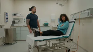 Een foto van presentator Serah die op een behandeltafel ligt. Er staat een behandelaar naast de tafel en kijkt naar haar. Serah houdt met haar linkerhand haar rechterelleboog vast.