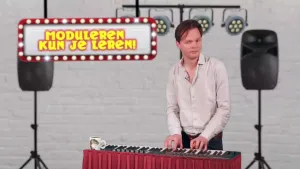 Een foto van een presentator die achter een elektrische piano staat en zijn handen op de toetsen van de piano heeft geplaatst. Daarnaast is de tekst "moduleren kun je leren!" zichtbaar. Op de achtergrond staan geluidsboxen en hangen podiumlichten.