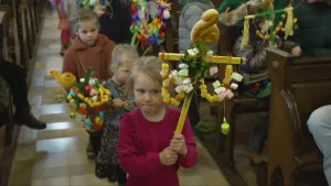 Een foto van een rij kinderen die in het gangpad tussen de kerkbanken lopen. De kinderen dragen een palmpasenstok in de vorm van een kruis. Op de stok staat een broodhaantje en aan de stok hangen naast een palmtak veel snoepjes.