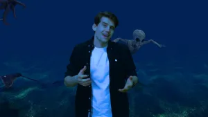 Een foto van een presentator met op de achtergrond de bodem van de oceaan. Naast de presentator zwemt een buitenaards wezen in de oceaan.  Aan de uiterste linkerkant zijn ook een deel van een octopus en rog zichtbaar. 