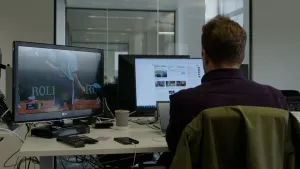 Een foto van een man achter een bureau met twee monitoren en een klein stukje van een laptopscherm zichtbaar. Op de linkermonitor is een tenniswedstrijd te zien en op de rechtermonitor een website van de krant De Telegraaf.