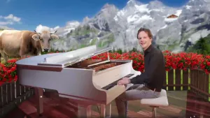 Een foto van een presentator achter een witte piano. De piano staat op een veranda met bloembakken met rode bloemen eromheen zichtbaar. Op de achtergrond staat een koe in een wei en zijn bergen met sneeuwtoppen te zien.