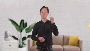 Een foto van een presentator die met zijn linkerhand een stemvork omhoog houdt. Op de achtergrond is een woonkamer te zien.