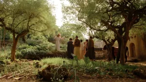 Een foto van Jezus die in een landschap met groene bomen en struiken een groep mensen toespreekt. Jezus staat op een iets hogere plek voor de mensen en de mensen kijken naar hem. Rechts naast de mensen zijn een ezel en de voorkant van een woning te zien.