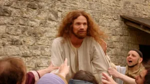 Een foto van Jezus die in het midden van de foto staat. Om Jezus heen staan allemaal mensen die met hun handen Jezus proberen aan te raken. Op de achtergrond is een stenen muur zichtbaar.