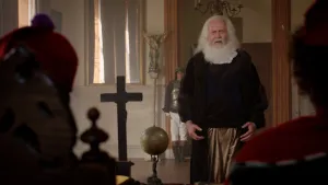 Een foto van Galileo Galilei die voor de inquisitie staat. Twee leden van de inquisitie zijn van de achterkant, zittend aan een tafel, zichtbaar. Op de tafel staan een houten kruis en een wereldbol. Op de achtergrond staat een beeld van een engel. 