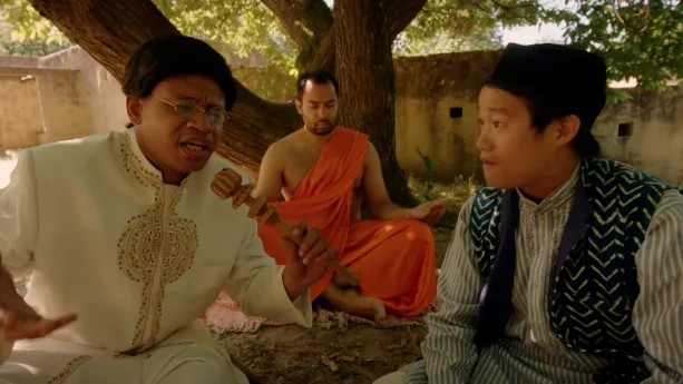 Een foto van Siddartha, ook wel de Boeddha genoemd, die onder een boom genaamd de Bodhiboom zit te mediteren. Op de voorgrond zit een presentator met een houten microfoon te praten met een dienaar van Siddartha, die naast hem zit.