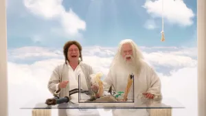Een foto van God en een Joods geklede interviewer die lachend aan een glazen tafel zitten in een blauwe wolkenlucht. Op de tafel staat een glazen aquarium met daarin een voorstelling van de schepping met Barbiepoppen die Adam en Eva voor moeten stellen.